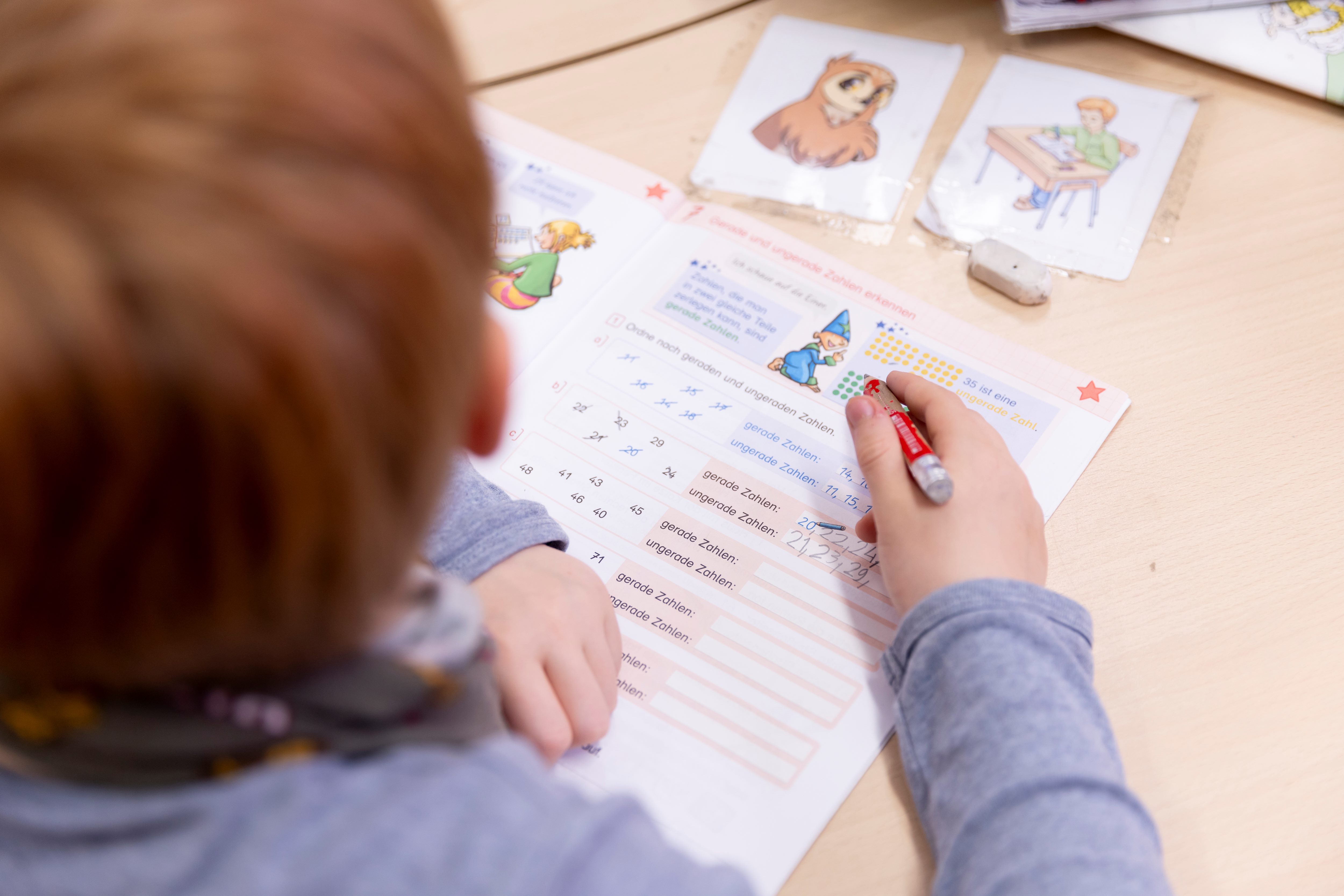 Die Kamera blückt über die Schulter eines Grundschülers im grauen Pullover. Das Kind hält einen roten Bleistift in der Hand und füllt ein Mathe-Arbeitsheft aus